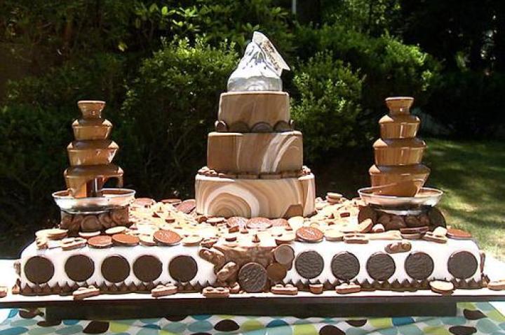 CHOCOLATE LOVERS CAKE
How was the cake made?
The cake was made of chocolate sponge cake (main cake), chocolate sponge cake (smaller cakes) with the following fillings: peanut butter cup filling, chocolate bar filling, peppermint filling, coconut flake and almond filling, marble chocolate fondant (covered the upper tiers), Hershey Kisses (border on bottom tier), melted Hershey Bars (chocolate fountain and river), Rice Krispies Treats (giant kiss on top of cake), and modeling chocolate (silver wrapper).
What's memorable about the cake?
Simeon's wife loves chocolate so he wanted Buddy to create the most "chocolately thing he could think of" that included her favorite chocolate bars. This cake was a chocoholic's dream complete with chocolate cake, candy bar fillings, a chocolate fountain and even a chocolate river!
Click "Next" to see the next sketch.