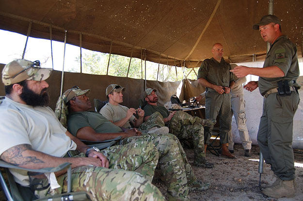 Oz (medic, Green Beret), Rob (intel, Navy SEAL ret.), Jeff "Biggs" (sniper, Navy SEAL fmr.), Craig "Saw" (team leader, Navy SEAL fmr.) all seated being briefed by Li Loeriet (standing with cap on head) from Quemic Anti-Poaching Unit about the situation of poaching in South Africa.