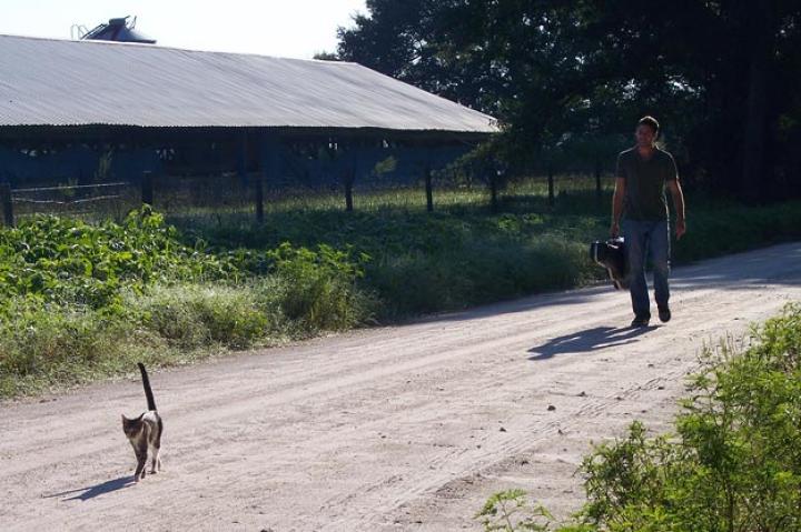 John travels along on dirt path with guitar and cat.