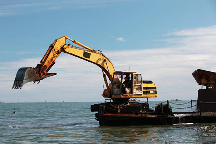 The barge's excavator crane extends out to dig up bottom sediment. A counterweight rig and stabilizing spikes held keep the craft level when the arm is extended.