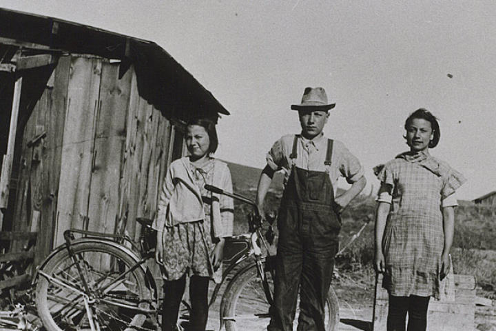 In this 1932 photograph, young John Schnabel and sisters Margaret and Mary stand in front of the shack they grew up in outside of Klamath Falls, Oregon. The 12 by 16 ft building was for the women while John and his brothers stayed in the chicken coop.