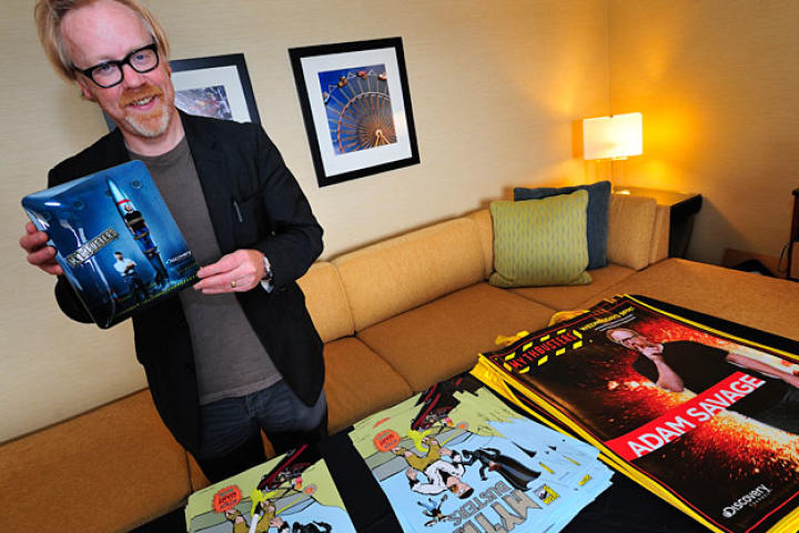 Adam Savage, flanked by the limited-edition posters and bags he autographed, shows off the custom MythBusters iPad that was rewarded to the fan who guessed his identity on the Comic Con floor.