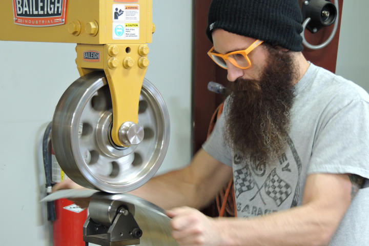 Aaron Kaufman, wearing orange glasses, runs sheet metal through a machine.