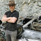 Adam Savage calmly gazes at the camera with his will-a-car-that-drives-over-a-cliff-really-explode-into-flames-upon-impact wreck in the background, in 2009.