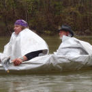 Our intrepid duo heads out in their latest ride, using the old tarp as both a boat skin and foul weather gear. Laying across Cody's lap is a jury-rigged paddle made from a forked branch wrapped with duct tape. The raft turns out to be fine in smooth water, but the floating scraps don't have much of a chance in the rapids that lay ahead.