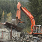 An excavator loads the shaker with a bucket of rock ore. Since this ore consists of weathered river sediments, most of the rocks — even the boulders — are rounded. Miners long ago realized that fewer edges makes the job of shakers and other processors more efficient. Below the shaker table the team installed a custom punch plate with 1 1/4 inch holes that only lets rocks smaller than this pass through to the next stage.