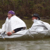 Our intrepid duo heads out in their latest ride, using the old tarp as both a boat skin and foul weather gear. Laying across Cody's lap is a jury-rigged paddle made from a forked branch wrapped with duct tape. The raft turns out to be fine in smooth water, but the floating scraps don't have much of a chance in the rapids that lay ahead.