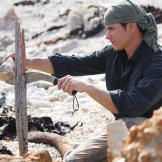 In Tasmania, Mykel uses a heavy rock as an assist while he splits a very hard piece of driftwood with his knife. Using even primitive tools, like this stone employed as a hammer, can save a lot of physical energy. Minimizing caloric expenditure is crucial when intake of food and water is so low.