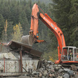An excavator loads the shaker with a bucket of rock ore. Since this ore consists of weathered river sediments, most of the rocks — even the boulders — are rounded. Miners long ago realized that fewer edges makes the job of shakers and other processors more efficient. Below the shaker table the team installed a custom punch plate with 1 1/4 inch holes that only lets rocks smaller than this pass through to the next stage.