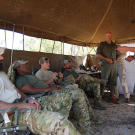 Oz (medic, Green Beret), Rob (intel, Navy SEAL ret.), Jeff "Biggs" (sniper, Navy SEAL fmr.), Craig "Saw" (team leader, Navy SEAL fmr.) all seated being briefed by Li Loeriet (standing with cap on head) from Quemic Anti-Poaching Unit about the situation of poaching in South Africa.